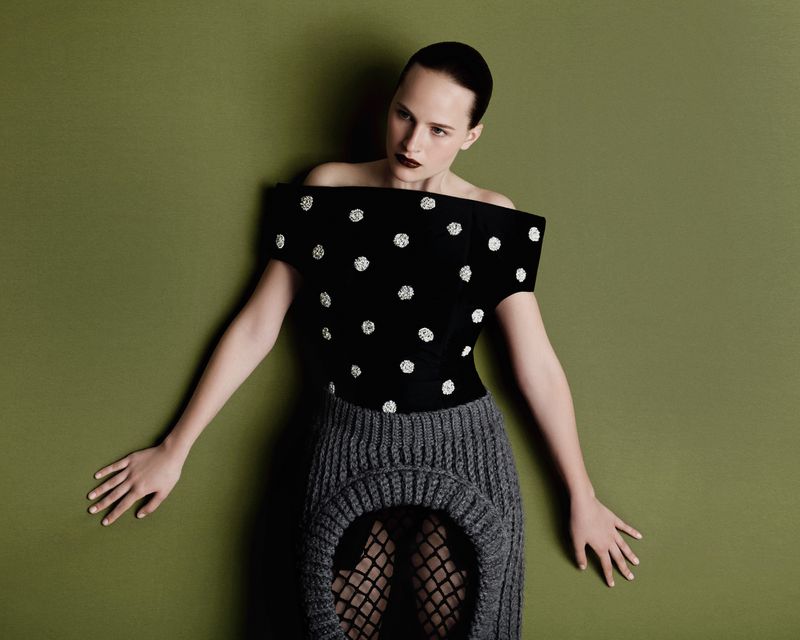 A woman with a shaved head poses against a green wall wearing a black and white polka-dot top and grey knit skirt in a studio shot.