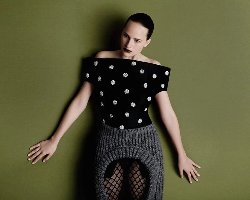 A woman with a shaved head poses against a green wall wearing a black and white polka-dot top and grey knit skirt in a studio shot.