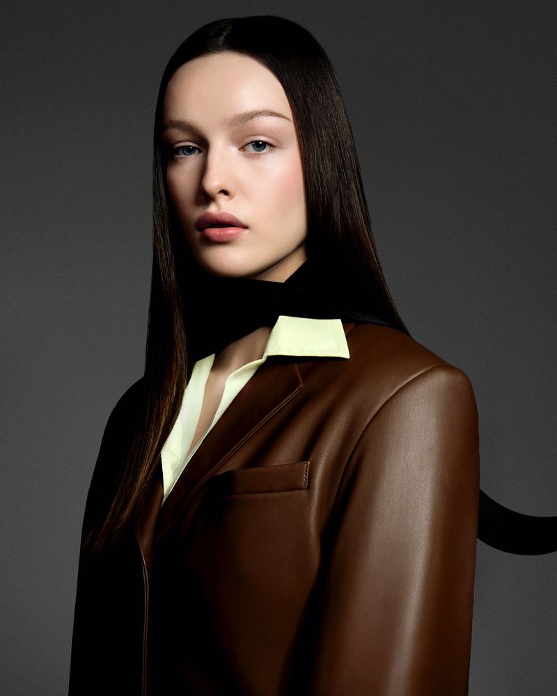 Portrait of a woman with dark hair wearing a brown blazer and light yellow top against a gray background in a studio setting.