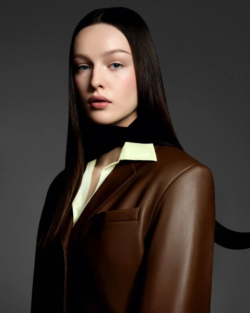 Portrait of a woman with dark hair wearing a brown blazer and light yellow top against a gray background in a studio setting.