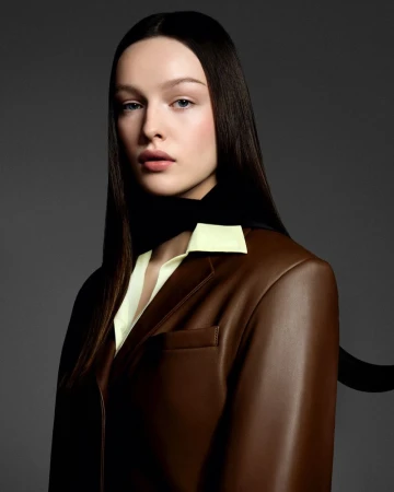 Portrait of a woman with dark hair wearing a brown blazer and light yellow top against a gray background in a studio setting.