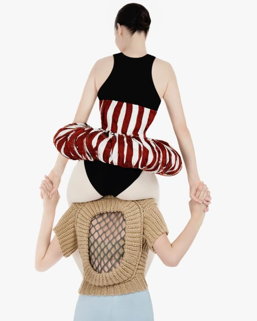 Two female fashion models are holding hands. One model is standing atop the other's shoulders, wearing a striped dress in a bright white studio.