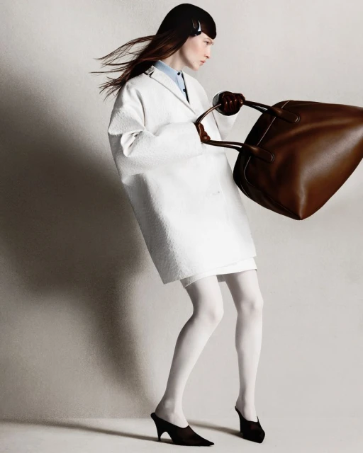 A woman in a white coat poses with a brown handbag in a studio setting, showcasing fashion and style with clean lines and neutral tones.