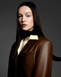 Portrait of a young woman with long dark hair wearing a brown leather blazer over a yellow shirt against a dark grey background