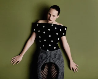 Model wearing a black top with white embellishments and a textured grey skirt against an olive green background.