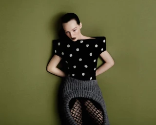 Model wearing a black top with white embellishments and a grey skirt against an olive green backdrop