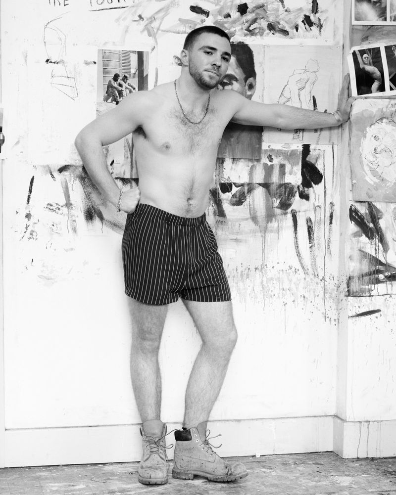 A shirtless man poses in striped shorts and work boots against a paint-splattered wall, creating an edgy, artistic, monochrome portrait.