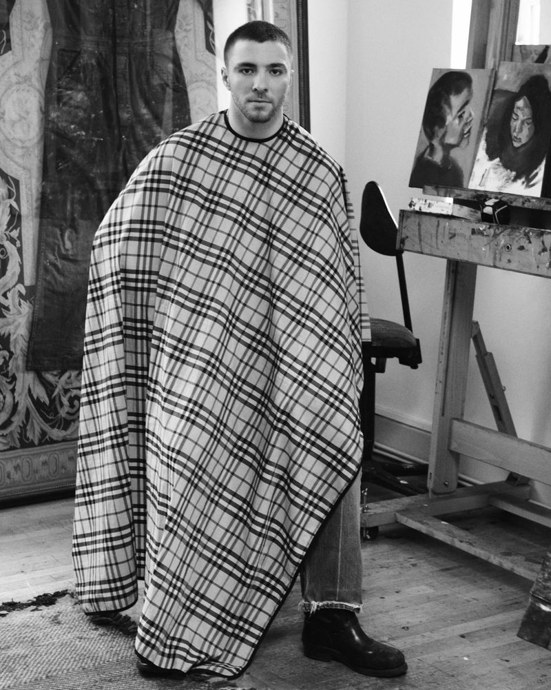 Monochrome portrait of a man in a plaid barber cape sitting indoors, with artwork and an easel visible in the background.