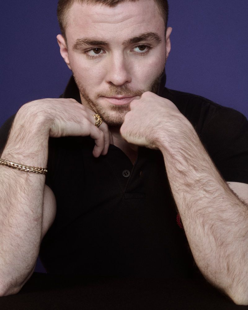 Portrait of Rocco Ritchie wearing a black polo shirt and gold bracelet against a solid blue background for Fantastic Man.