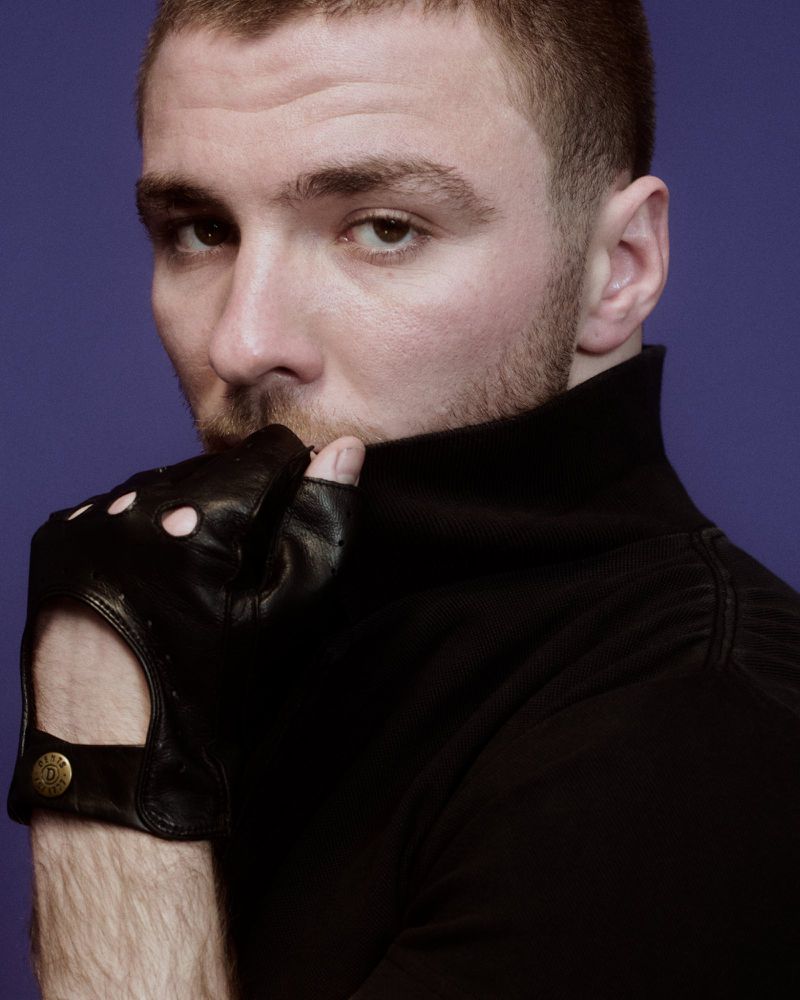 Close-up studio portrait of a young adult man in a black turtleneck sweater and fingerless leather glove against a purple background.