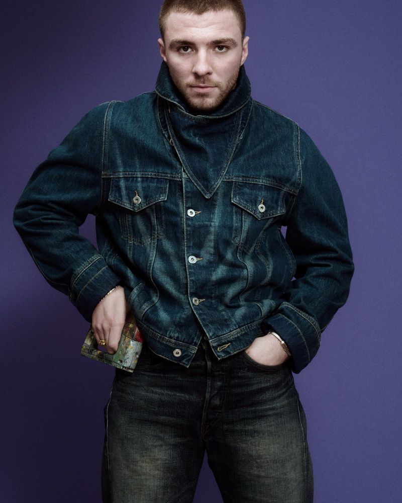 Portrait of a handsome man wearing a denim jacket and jeans against a purple backdrop, looking directly at the camera.