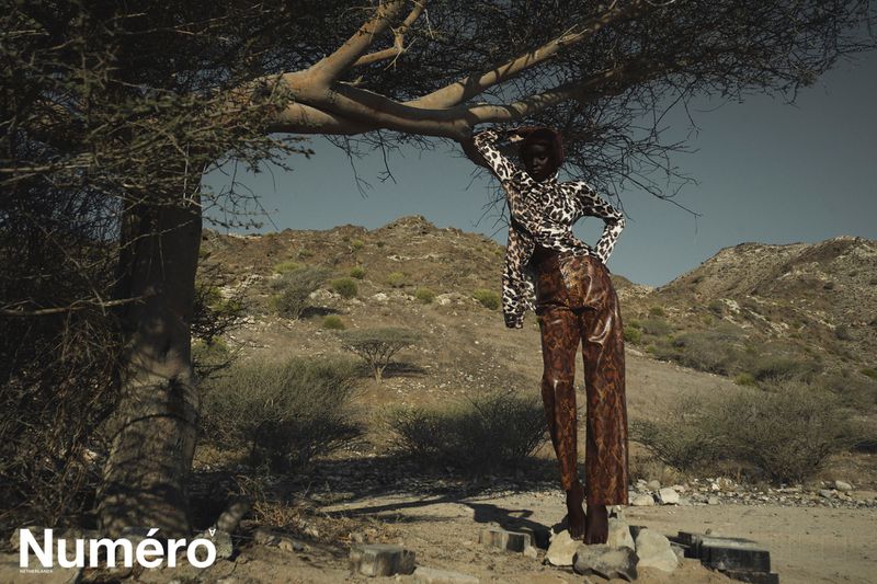 A dark-skinned fashion model in a patterned outfit poses under a tree against a desert mountain backdrop in a fashion editorial photo.