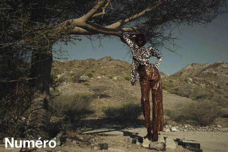 A dark-skinned fashion model in a patterned outfit poses under a tree against a desert mountain backdrop in a fashion editorial photo.