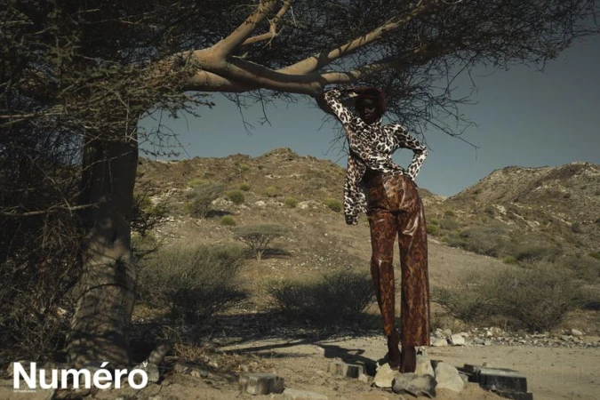 A dark-skinned fashion model in a patterned outfit poses under a tree against a desert mountain backdrop in a fashion editorial photo.