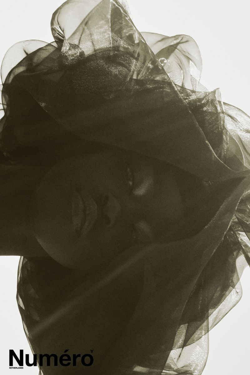 Close-up portrait of an adult female with sheer black fabric draped over her head against a bright backdrop in a beauty editorial.