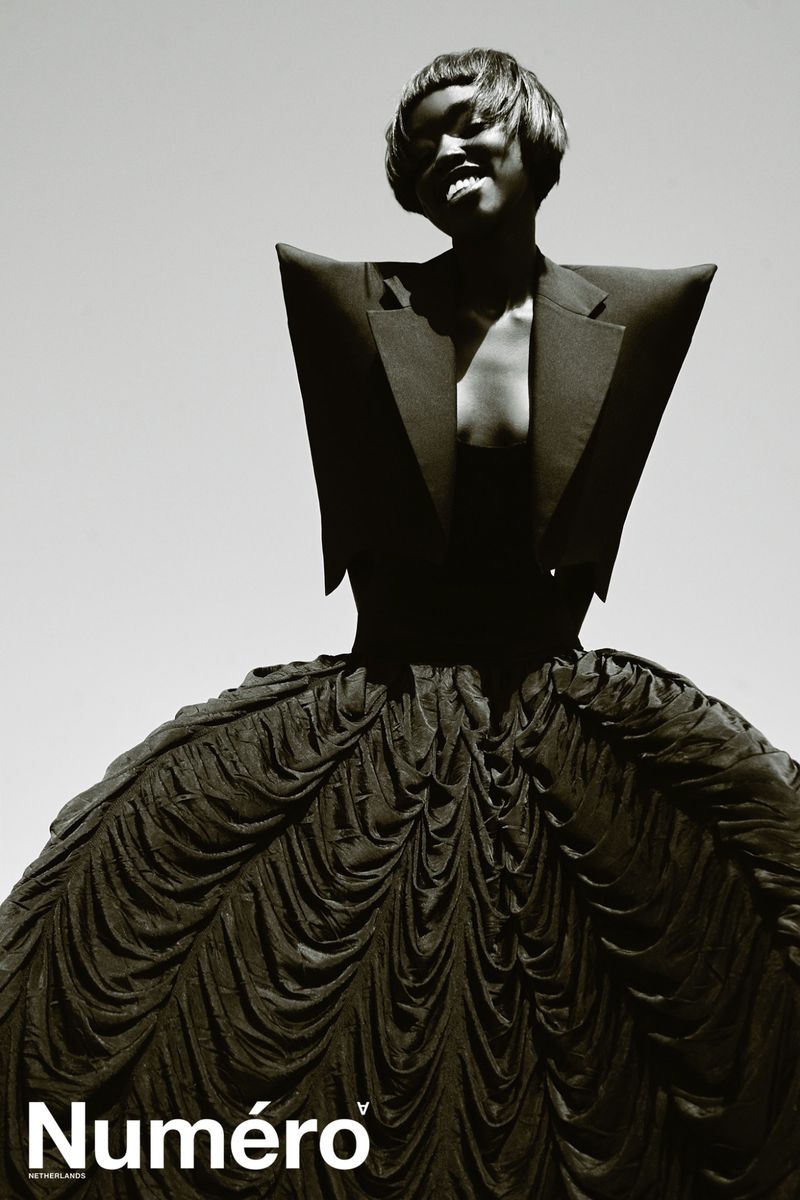 Fashion editorial shot of a Black woman posing in a sculptural black dress and jacket, set against a bright sky, magazine logo visible.