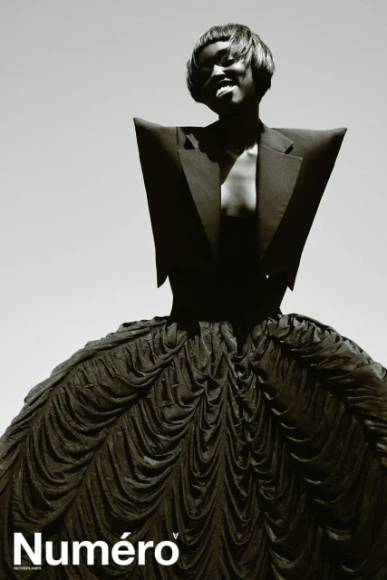 Fashion editorial shot of a Black woman posing in a sculptural black dress and jacket, set against a bright sky, magazine logo visible.