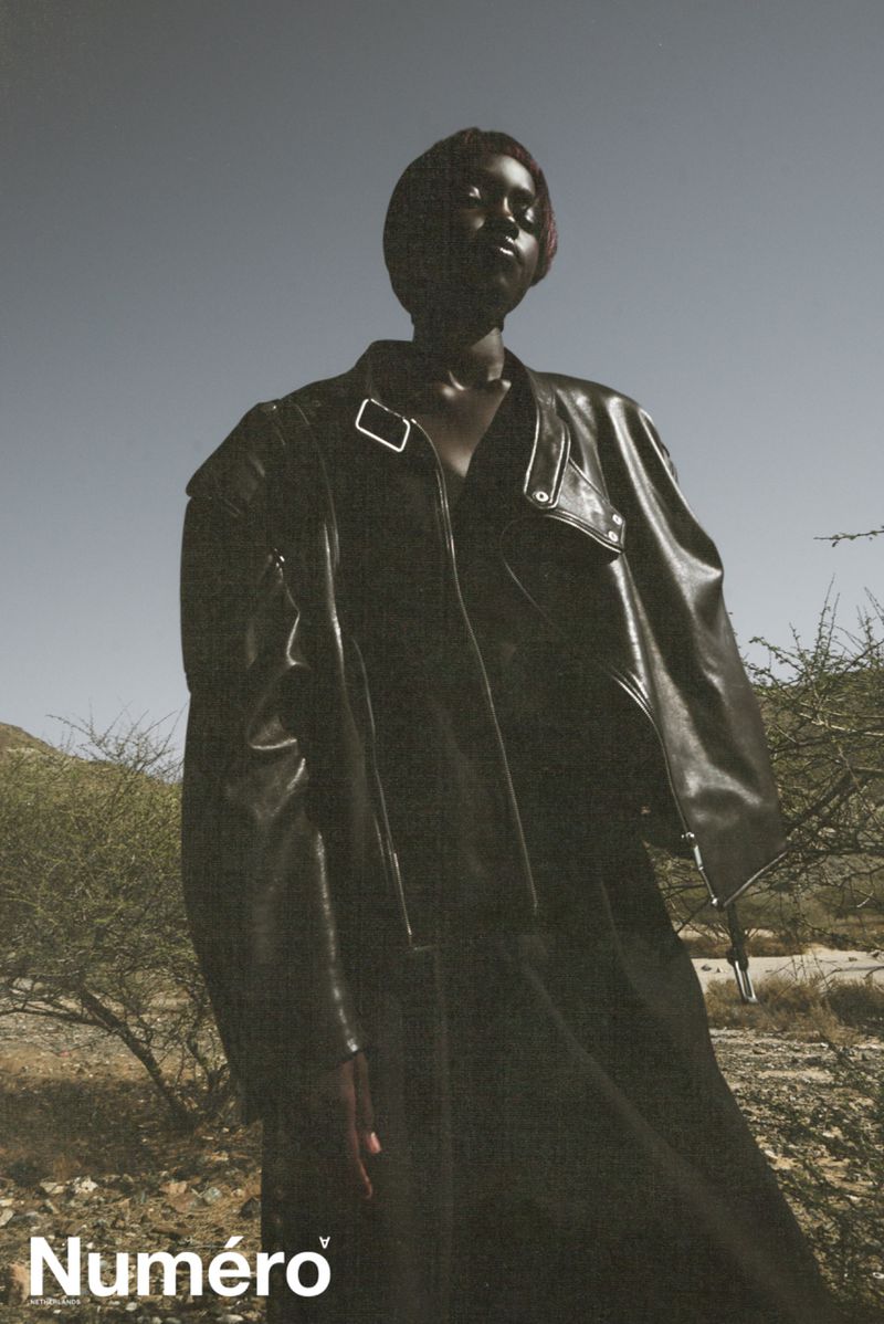 Fashion shot of a Black model in a black leather jacket and dress, posing outdoors against a clear sky in an arid landscape.