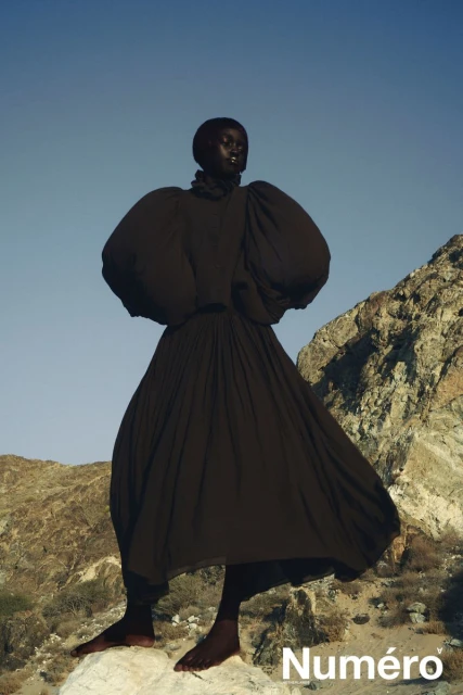 Editorial fashion shot of a Black female model in a dramatic black dress standing on a rock formation against a clear blue sky.