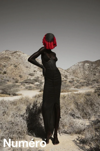 Editorial fashion shot of a Black female model posing in a black dress and ornamental headwear, against a desert and mountain background.