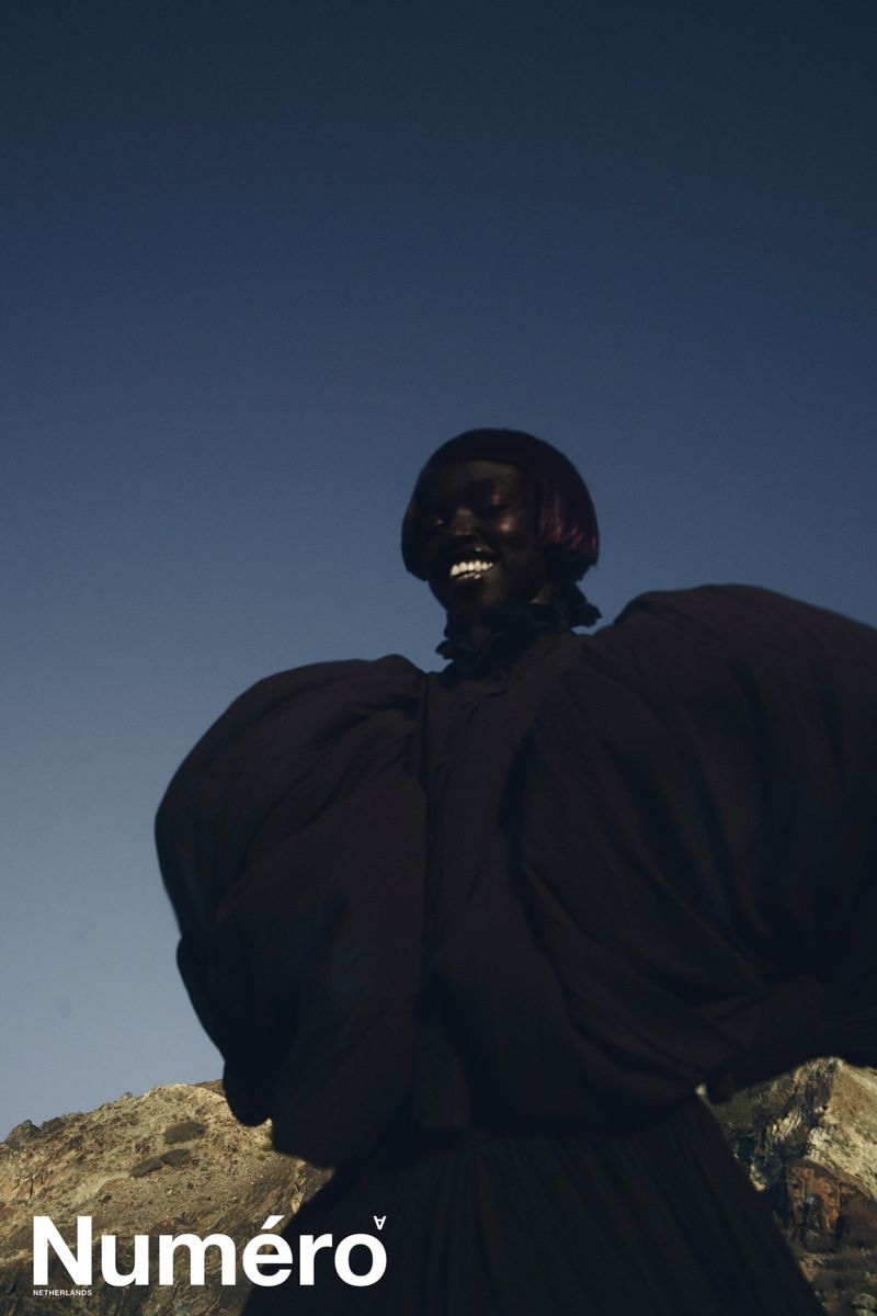 A smiling Black woman in a dark dress with large puffy sleeves poses against a blue sky and mountain range in an outdoor fashion shot.
