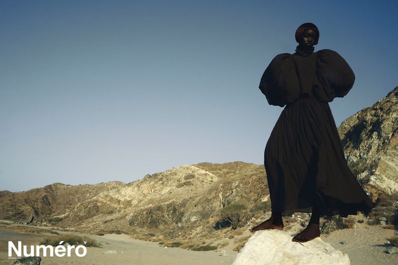 A dark-skinned woman in a black dress stands on a rock formation against a desert mountain landscape and clear blue sky in an editorial fashion shot.