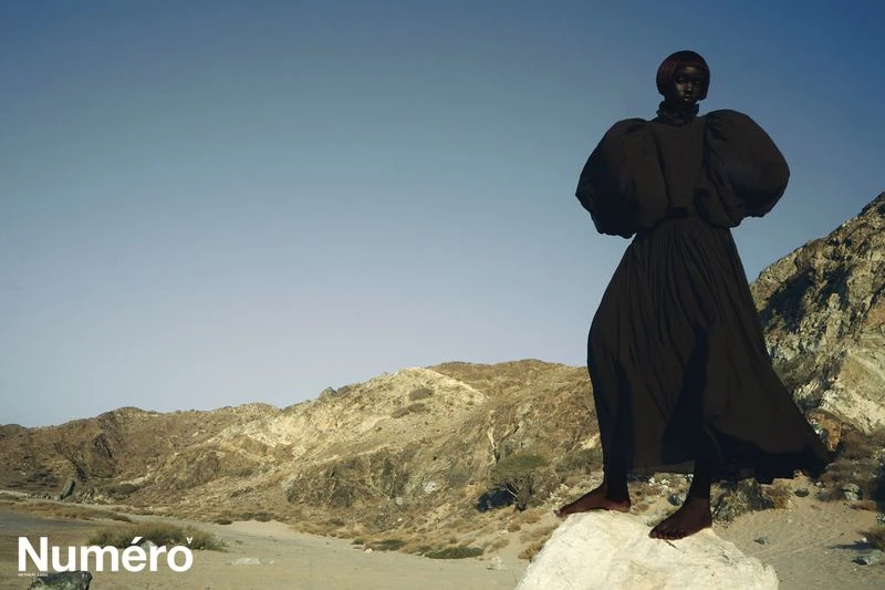 A dark-skinned woman in a black dress stands on a rock formation against a desert mountain landscape and clear blue sky in an editorial fashion shot.