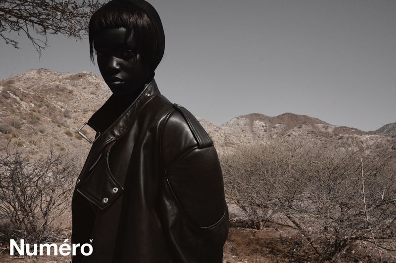 Woman in black leather jacket poses against a desert mountain backdrop. Fashion shot with overcast lighting and muted color palette.