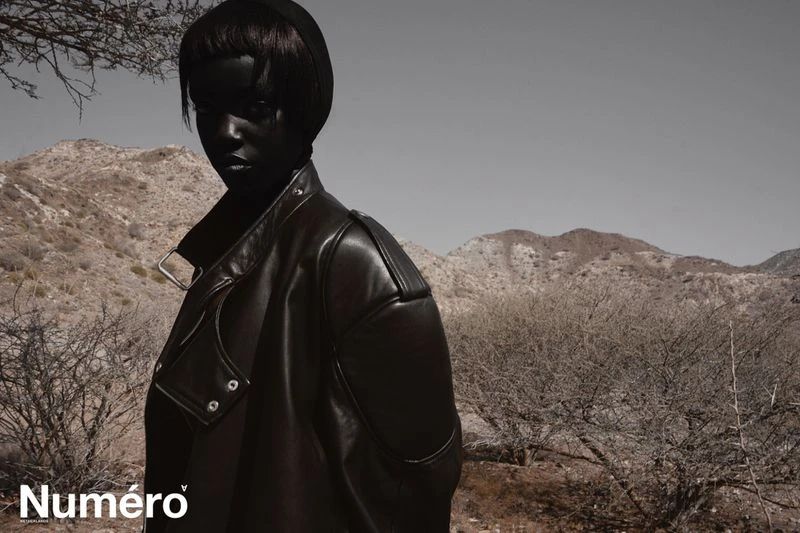 Woman in black leather jacket poses against a desert mountain backdrop. Fashion shot with overcast lighting and muted color palette.