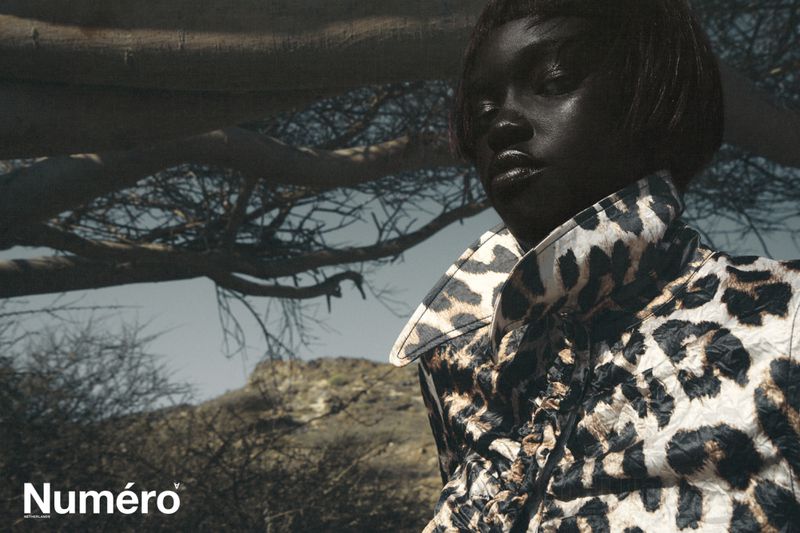 Fashion editorial shot of a Black woman wearing a leopard print jacket, posing in a landscape with trees and a distant hill.