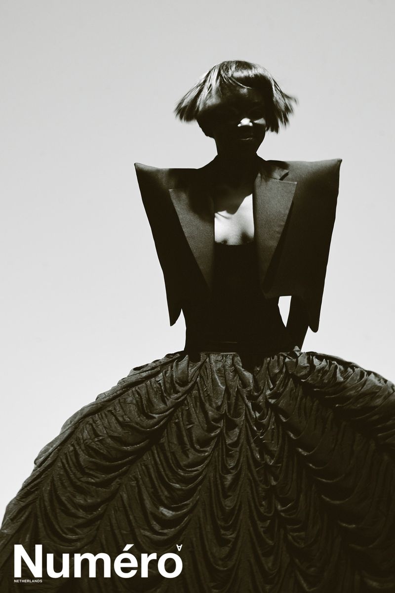 Editorial fashion portrait of a woman with a dark bowl cut wearing a black gown, dramatically lit against a bright background.