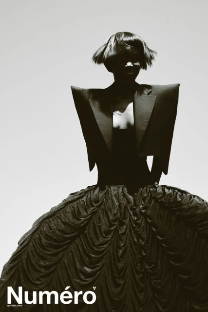Editorial fashion portrait of a woman with a dark bowl cut wearing a black gown, dramatically lit against a bright background.