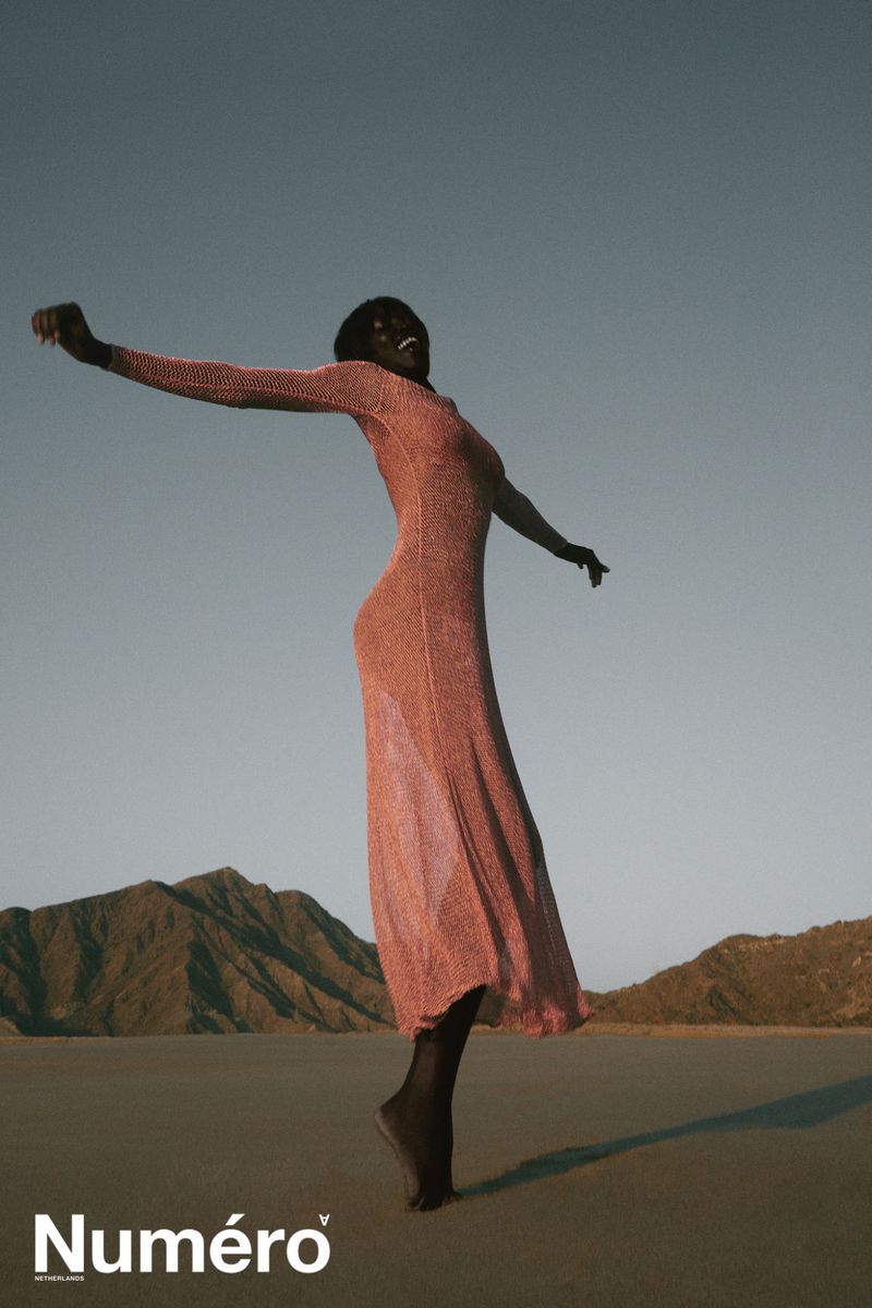 An adult woman poses in a long rose colored dress on a beach at dusk with mountains in the background under a blue and rose colored sky.