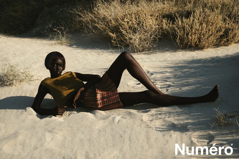 An African-American fashion model is lying on her side in beach sand wearing a mustard-colored top and patterned skirt in a desert landscape.
