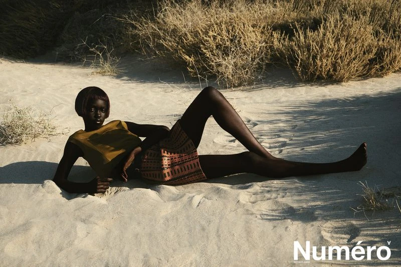 An African-American fashion model is lying on her side in beach sand wearing a mustard-colored top and patterned skirt in a desert landscape.