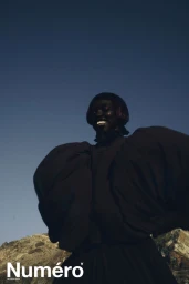 Model Alek Deng wearing dark voluminous clothing against a mountain landscape and clear blue sky for Numero Netherlands editorial.