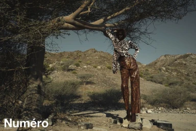 Model Alek Deng wearing a leopard print top and brown patterned trousers leaning against a tree in a desert landscape for Numero Netherlands.