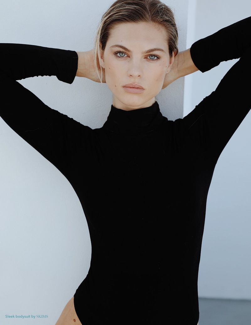 Blonde woman in black turtleneck bodysuit poses with her hands behind her head against a white background in a fashion studio shot.
