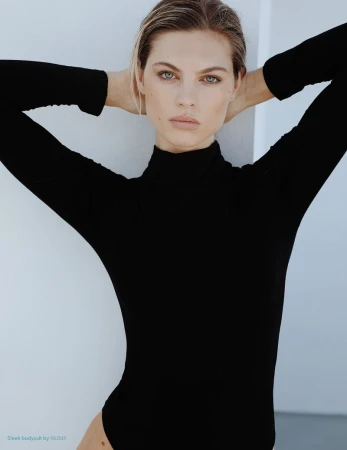 Blonde woman in black turtleneck bodysuit poses with her hands behind her head against a white background in a fashion studio shot.
