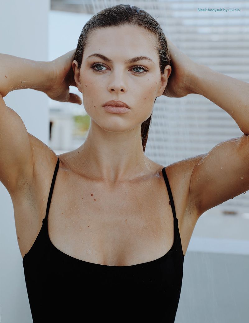 Close-up studio portrait of a wet adult woman in a black swimsuit with her hands behind her head, looking off to the side.