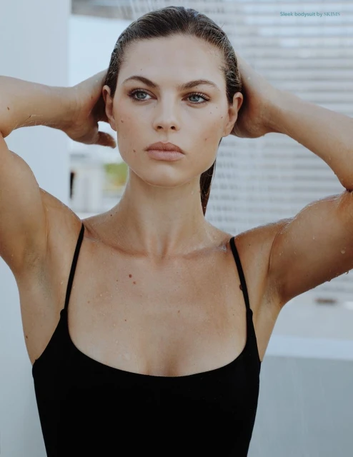 Close-up studio portrait of a wet adult woman in a black swimsuit with her hands behind her head, looking off to the side.