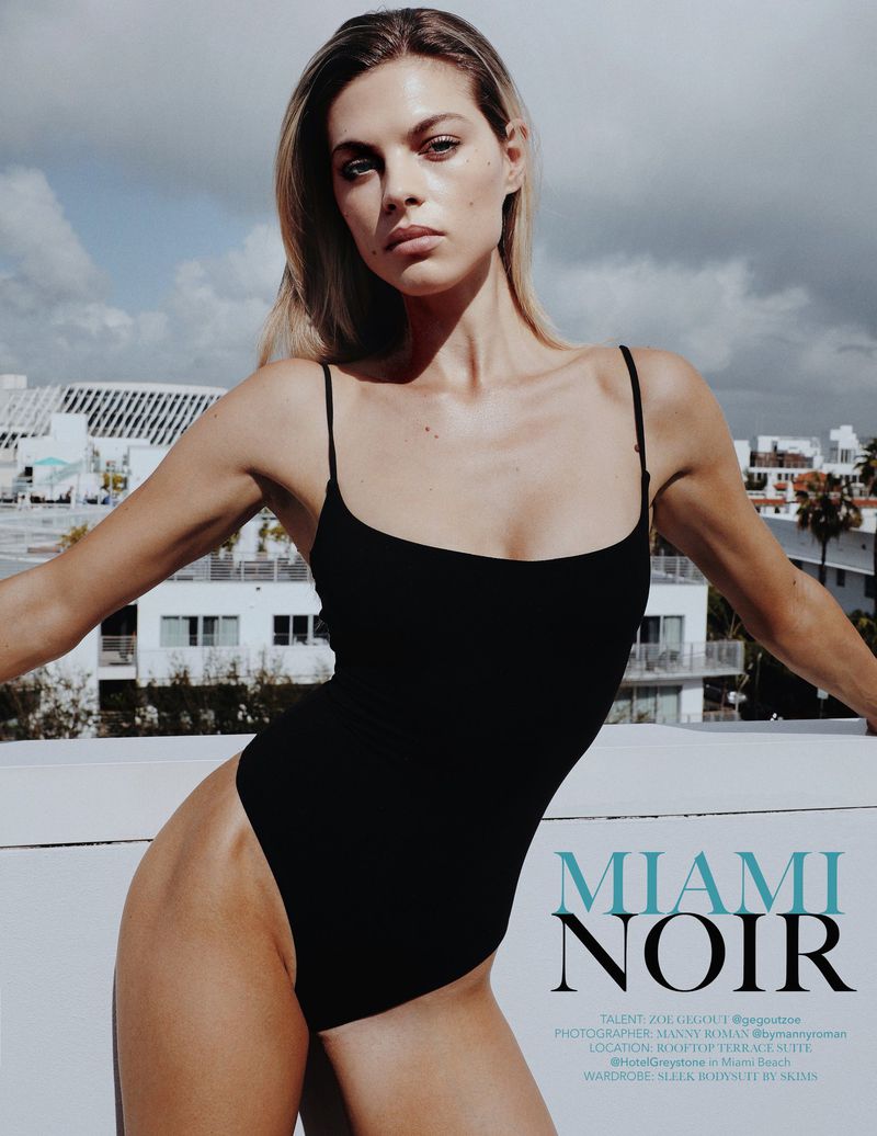 Woman in a black bathing suit poses on a rooftop with city buildings in the background under a cloudy sky in a fashion shot.