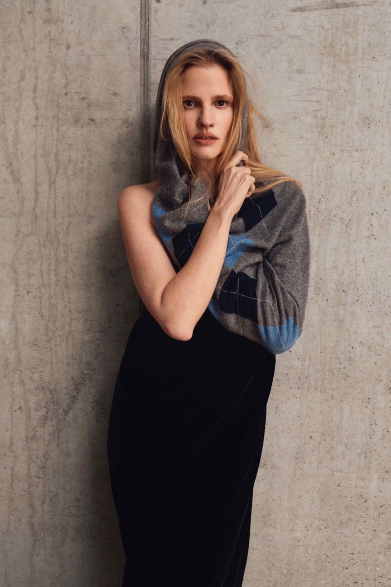 Indoor studio shot of a woman posing in a black skirt and gray argyle top with a hood. She is posing against a concrete wall.