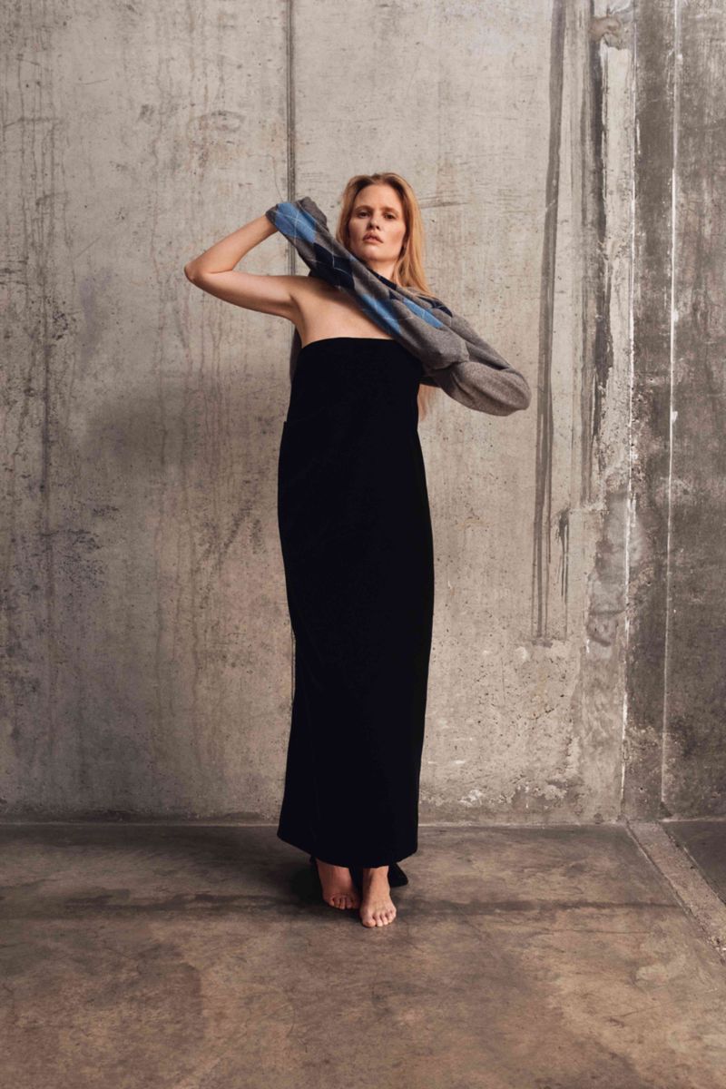 Blonde woman poses in a long black dress in front of a concrete wall, holding a blue and gray patterned sweater on her shoulder.