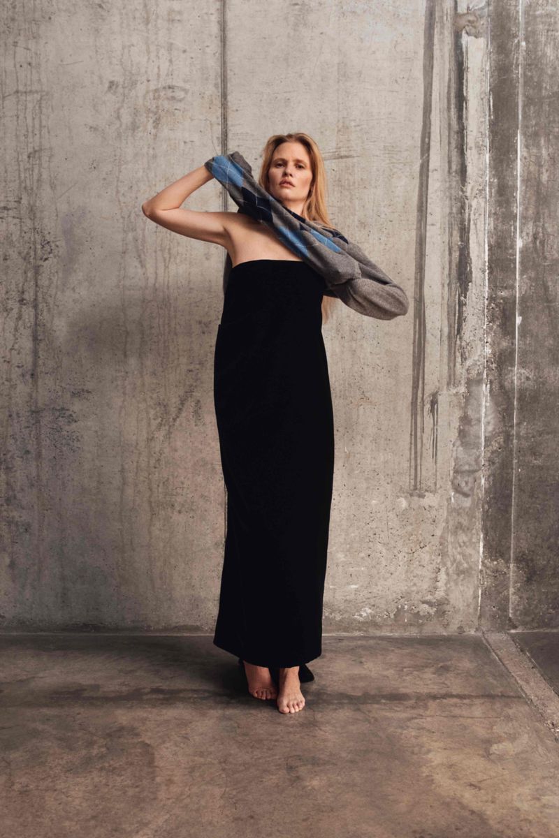 Woman in long black dress stands in minimalist setting, holding gray and blue jacket, looking at camera. Neutral color palette.