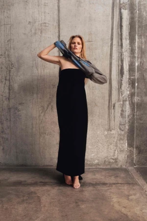 Woman in long black dress stands in minimalist setting, holding gray and blue jacket, looking at camera. Neutral color palette.