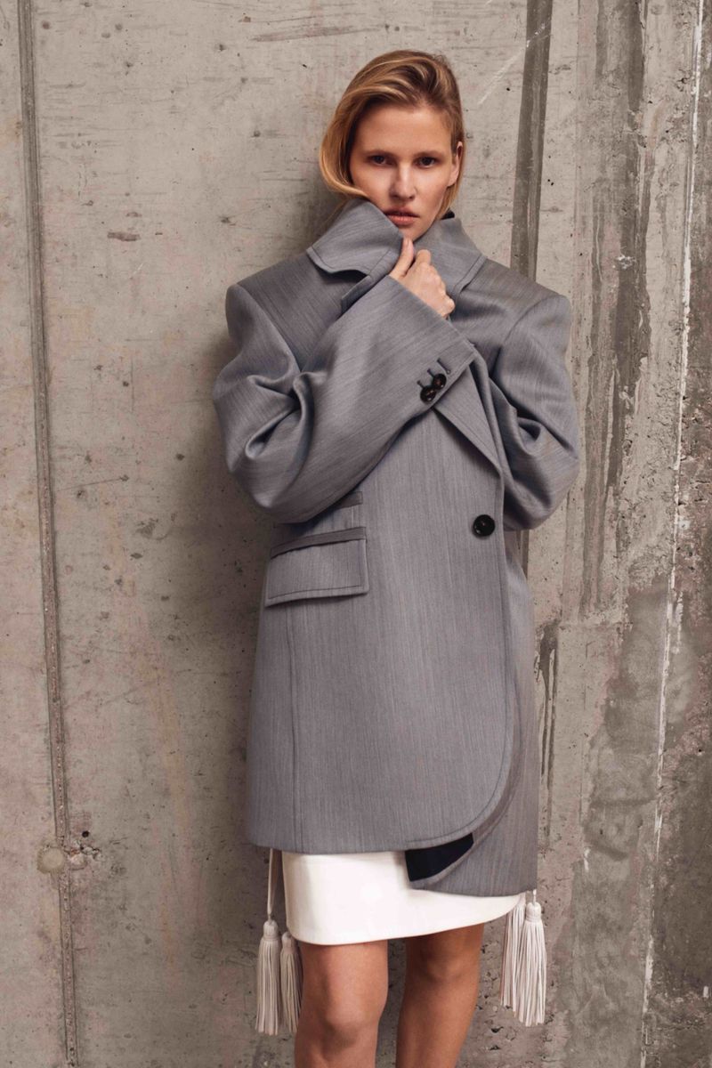 Fashion photo of a woman wearing a gray oversized blazer with tassels over a white dress, posing against a concrete wall.