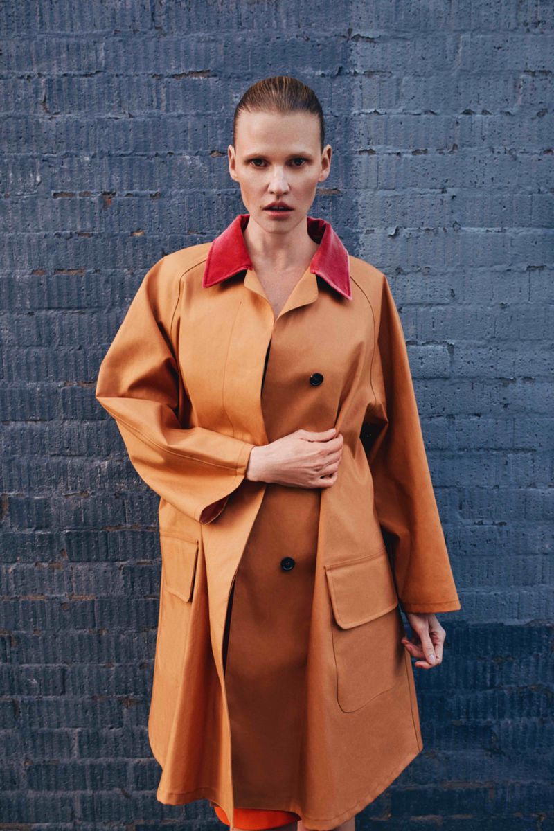 A woman in a tan trench coat with a red collar posing outdoors against a textured blue brick wall in a fashion editorial style.