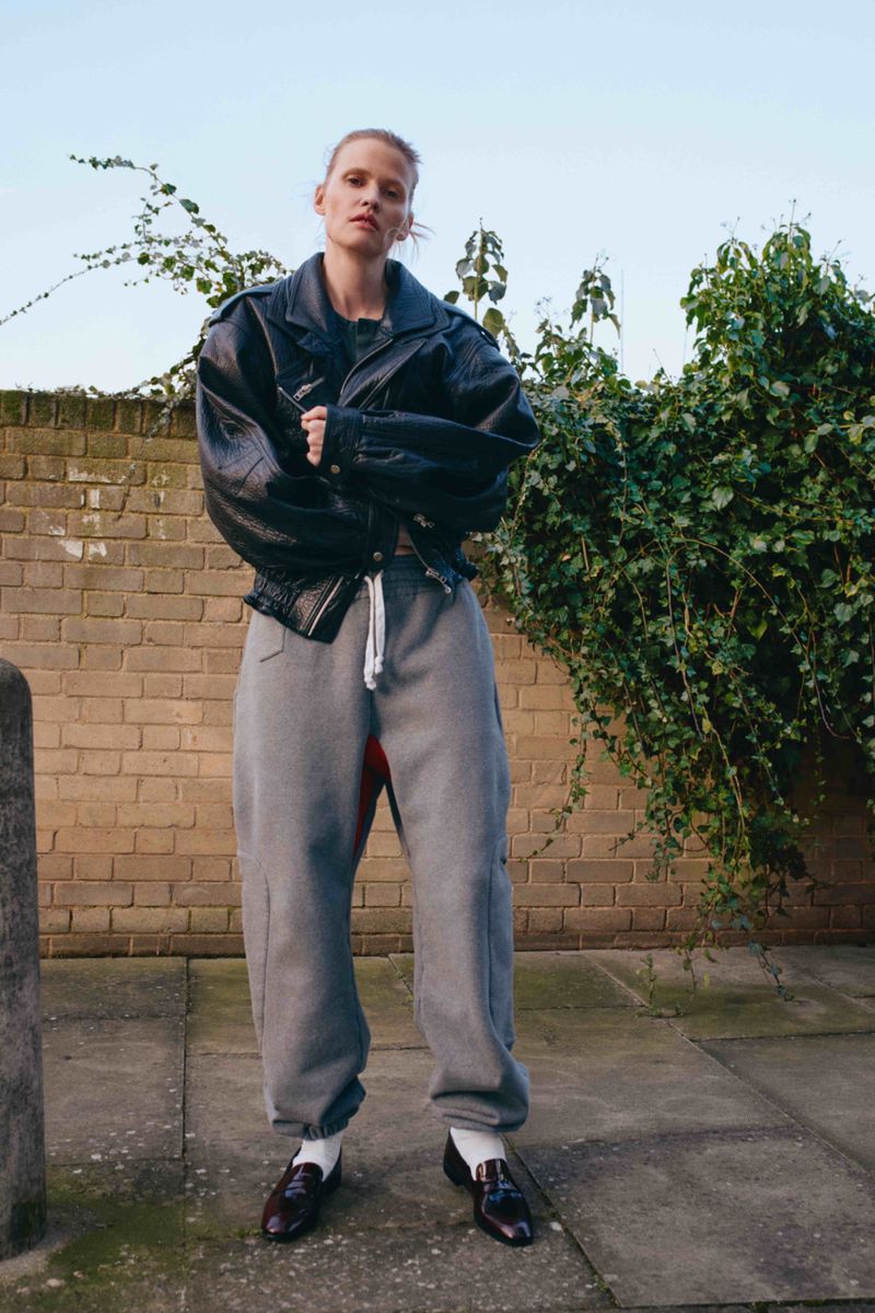 A woman stands outdoors, wearing a black leather jacket, gray sweatpants, white socks, and loafers, with a brick wall behind her.