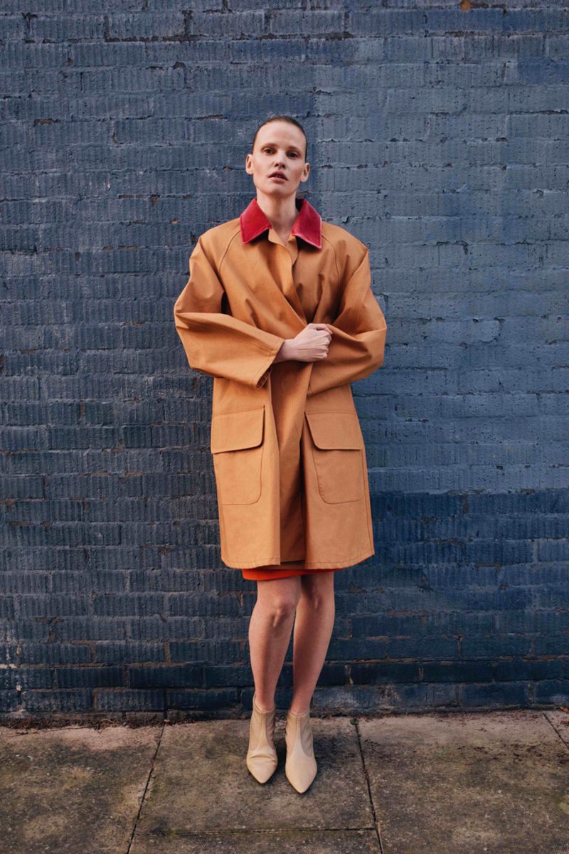 A woman in a tan trench coat stands against a textured blue brick wall, complemented by beige boots and a pop of red at the collar.